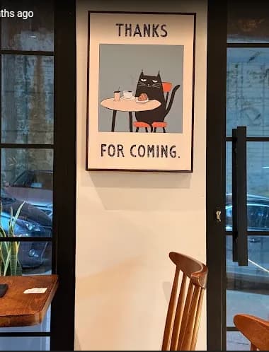 Une affiche encadrée près de la porte du café : un chat noir attablé devant un café et une pâtisserie, au-dessus des mots Thanks for coming.