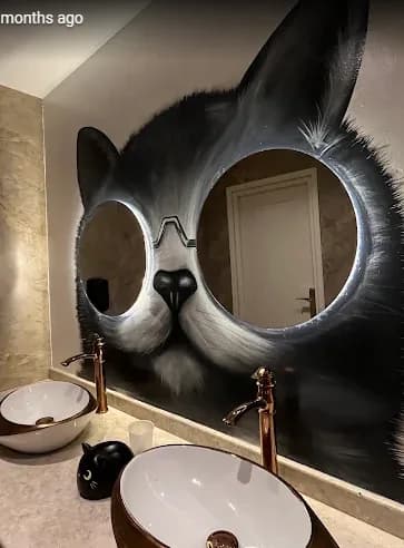 Un mur de toilettes peint d'une tête de chat noir et blanc surdimensionnée, dont un miroir rond forme l'œil, au-dessus d'une vasque en pierre.